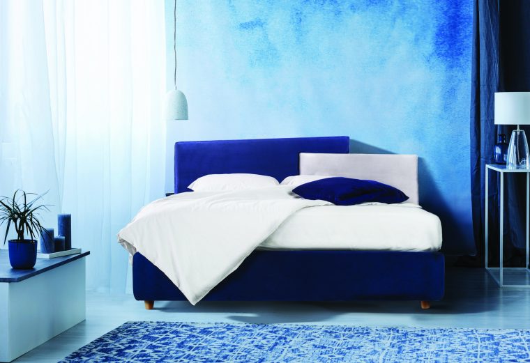 Grey boxes next to bed with blue bedding in bedroom interior with white lamp against a curtain